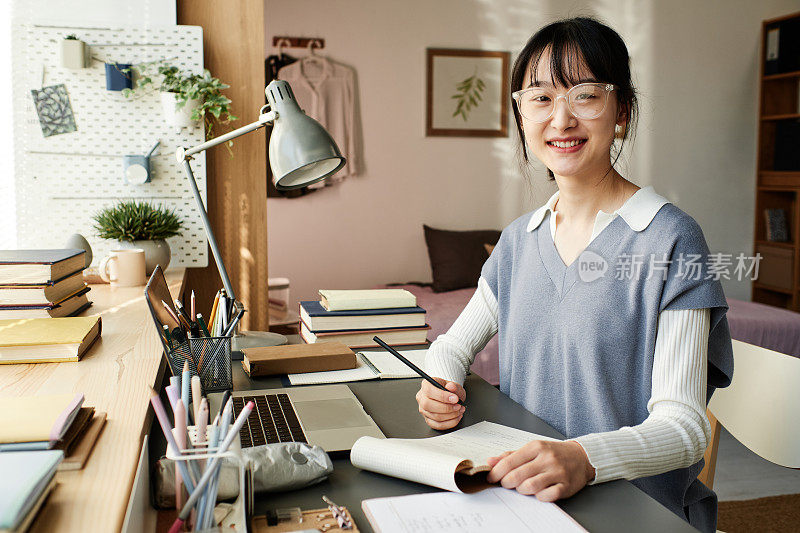Portrait of happy Asian student girl in transparent glasses sitting at desk with laptop and making notes in sketchpad